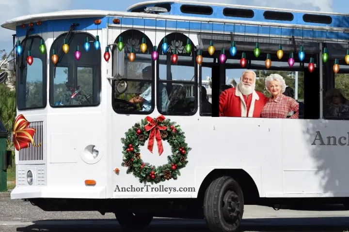 Holiday-themed trolley decorated with lights and wreath, carrying Santa and a woman.